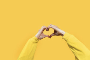 A hand is gesturing a love heart symbol against a yellow wall to demonstrate inclusion and engagement 